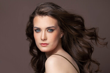 Naklejka premium Portrait of beautiful young woman with blowing long healthy dark hair and clear skin on brown background
