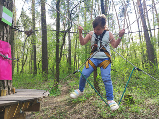 Little Caucasian blond girl plays in the park or forest, rope park in outdoors. Summer sports...