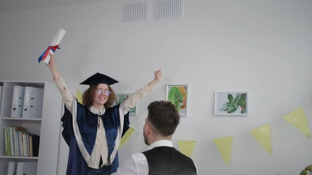 girl in mantle and hat jumps on couch and hugs male friend rejoices at graduation from institute and certificate of education