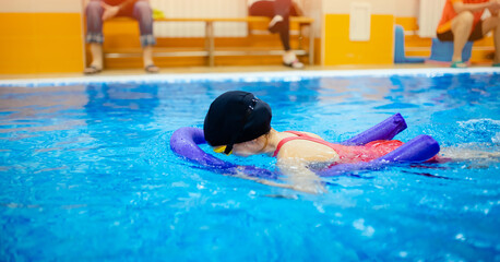 Child girl is engaged in swimming with trainer in pool, uses polyurethane stick to practice stroke