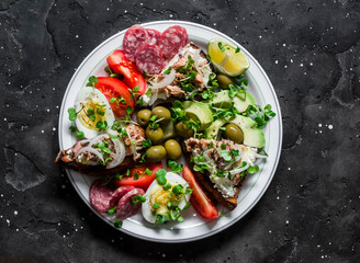 Large snack, appetizers, tapas plate - salami, mackerel cream cheese sandwiches, olives, avocado, tomatoes on a dark background, top view