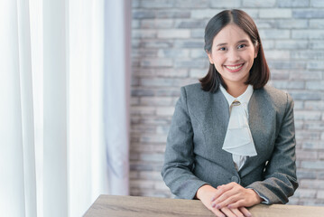 Beautiful Asian business woman secretary receptionist wait for client with happy friendly smile. Manager prepare for meeting, sitting in indoor office put hands on table in professional polite manner