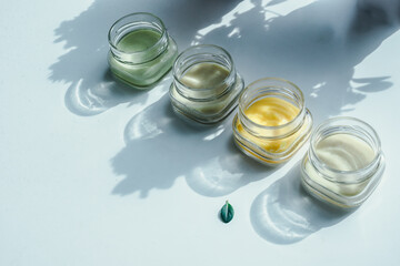Four jars of cosmetic cream stay on the white surface in a shadow of flowers with green leaf in front of them. Pastel colors, herbal cosmetics concept.