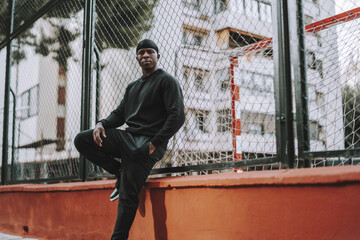 Confident Black Spanish male in black casual clothes and do-rag leaning on a wire fence