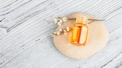 Glass bottle with essential oil and a sprig of a flower lying on a stone on a wooden white background, top view, copy space. Aromatherapy, massage oil, natural organic cosmetic, alternative medicine.