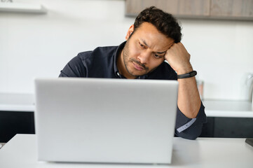 Upset multi-ethnic man is using laptop sitting at home alone, pensive young indian guy sits in the kitchen at with the laptop, rested head with hand, has financial problems and issues with project