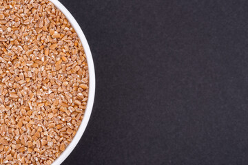White glass bowl of wheat groats. Isolated on a dark grey background. Top view close up