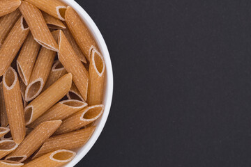 White glass bowl of raw brown penne rigate pasta. Isolated on a dark grey background. Top view close up