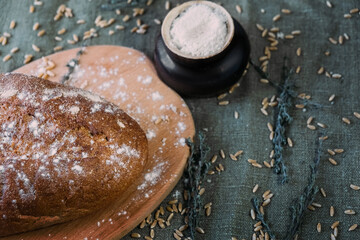 Fresh multigrain bread sprinkled with flour on a wooden board. Around the grain of wheat and grass. Nearby is a pot of salt. View from above.