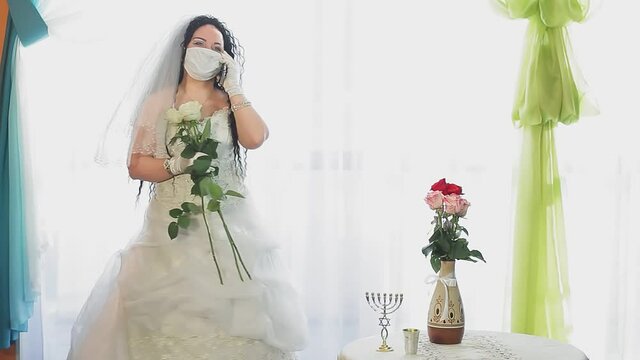 A Jewish bride in a wedding dress and veil wearing a medical mask in a synagogue hall before the chupah ceremony speaks on the phone