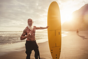 Senior man having fun surfing during sunset time - Retired male training with surfboard on the beach - Elderly healthy people lifestyle and extreme sport concept - obrazy, fototapety, plakaty