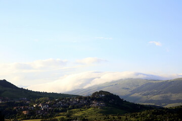 View of village perched on the mountains, among green trees, and other mountain topped by clouds in the background, Abruzzo, Italy