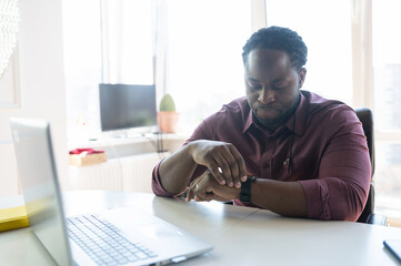 Confident African-American businessman looks at the smart watch wearing on his wrist, using laptop sitting at the desk indoor, checks messages or schedule using a trendy device