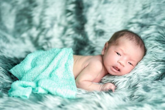 The newborn is wrapped in a green cloth, lying face down on a soft, gray rug. She is making a innocent face. Soft focus