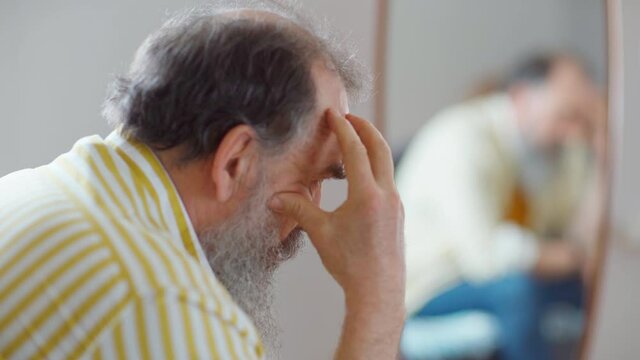 Side view head and shoulders shot of depressed pensive senior man with gray beard rubbing his face sitting near mirror at home or in hotel, feeling frustrated and thinking what to do