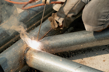 A welder is sitting on a steel roof frame and is welding steel pipes and wearing masks and leather gloves.