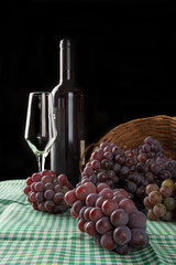 Grapes, basket and overturned box with grapes falling on the table along with wine and a glass on a checkered tablecloth, black background, selective focus.