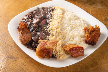Brazilian feijoada, white dish with a delicious Brazilian feijoada, rice, farofa, and fried pork with two oranges on rustic wood, selective focus.