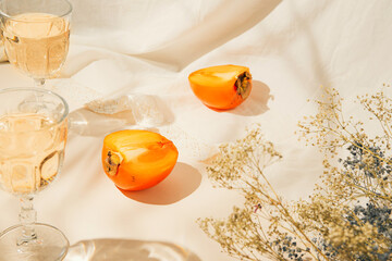 Halfs of persimmon fruits and two glasses with lemonade on pastel background with white cloth and sunlit. Summer drinks and refreshment concept. Minimal style composition.