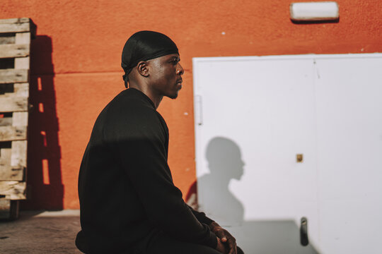 Pensive Black Spanish male in black clothing and do-rag posing outside of a building