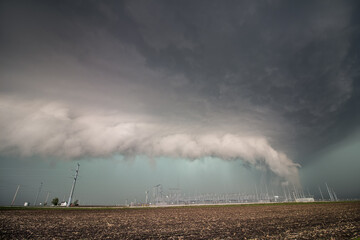 A severe thunderstorm and low shelf cloud loom over electrical equipment and power poles.