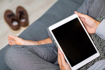 Man sitting on the sofa with a tablet on his lap. Barefoot man with shoes in the background. 