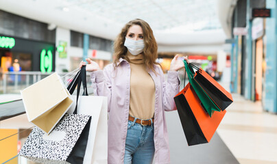 Portrait of a young woman shopaholic wearing medical mask in a shopping mall. A woman holds a lot of bags with clothes from different stores. Shopping during coronavirus Covid-19 lockdown