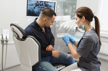 Young woman dentist selecting a shade of enamel for implantation. Male patient consults on dental treatment in a modern dental clinic
