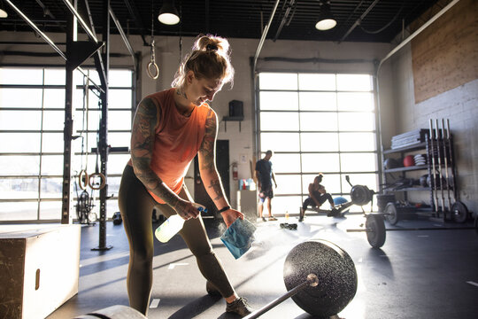 Woman disinfecting barbell with spray cleaner and cloth in sunny gym