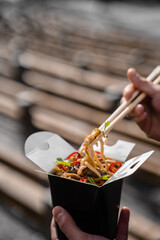 Wok in box in black food container. Holding spicy noodles with chopsticks. Fast food delivery service. Takeaway chinese street meal.
