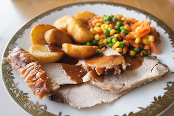 Home cooked pork roast dinner with crackling, gravy, roast potatoes and vegetables on a white plate with a gold trim.
