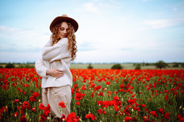Beautiful girl posing in a poppy field. Young woman stands in a spring blooming garden. A curly-haired girl stands in blooming field in summer. Sunny day, summer landscape. Joy and relaxation concept.