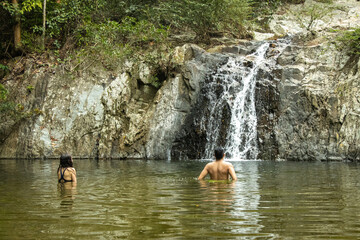 Pareja con el agua hasta la cintura caminando por un rio en medio de la naturaleza junto a una cascada