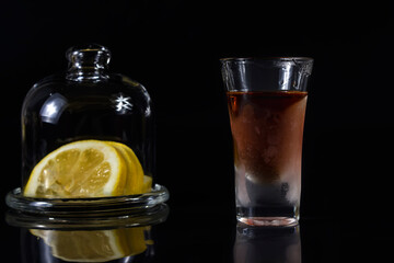 A glass of cognac on a black background next to a lemon in a container. Close-up