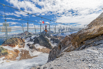 Woman on suspension bridge waving a Swiss flag. Titlis cliff walk with glacier in the Uri Alps. Viewpoint at 3028 meters in Summer season, Bern canton. The highest suspension bridge in Europe. - obrazy, fototapety, plakaty