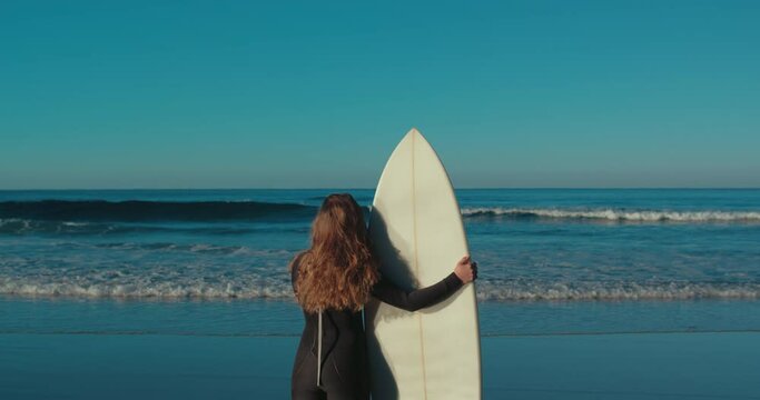 Back view of surfer female figure stand on edge of waves and sandy beach hold surfboard and look over horizon. Cinematic nomad vanlife lifestyle of surf affectionate. Woman in moment of peace and calm