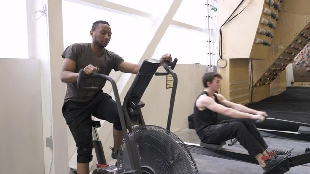 Young men exercising on elliptical bike and rowing machine in gym