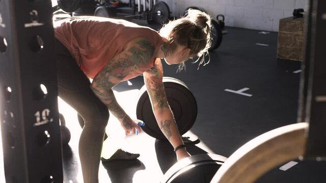 Young woman with tattoos disinfecting barbell with cleaner in gym