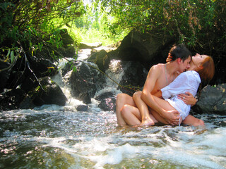 Young loving couple sits in the water near the waterfall. Guy with a girl passionately hugging near the waterfall.