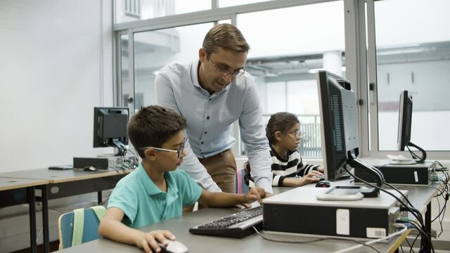 Cheerful teacher and schoolboy working on computer together. Smiling teacher typing on keyboard, schoolkid in eyeglasses sitting and repeating after him. Computer science, education concept. 