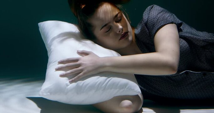 Cinematic close-up, young beautiful woman in pyjamas sinking under water, head on pillow, looking at camera slow motion.