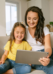 Fototapeta na wymiar Young loving mother with cute little daughter looking at digital tablet and smiling, having video chat with family while spending leisure time tigether at home. Kids and modern technologies concept