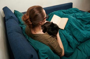 photo from the back of a woman reading a book with a cat in her arms, education at home, a hobby