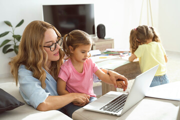 Young mom woman trying to work on laptop from home while her two little kids daughters playing...