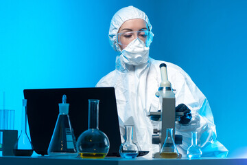 A chemist studies substances under a microscope and records the data in a laptop. A scientist in a chemical laboratory. A lab technician in a protective suit and glasses.