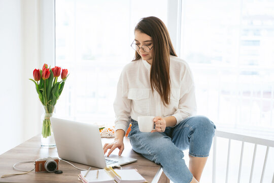 Young woman blogger working remotely on laptop while sitting on table near window at home, female photographer choosing photos on computer and drinking coffee. Distance work and freelance concept