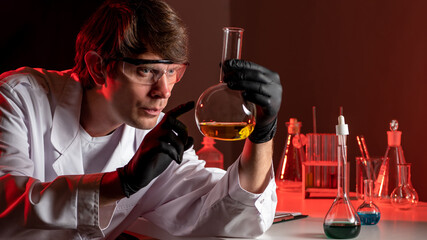 A chemist wearing safety glasses on a red background. A chemist examines a chemical liquid in a flask. Scientific research in chemistry. A scientist in a chemical laboratory.