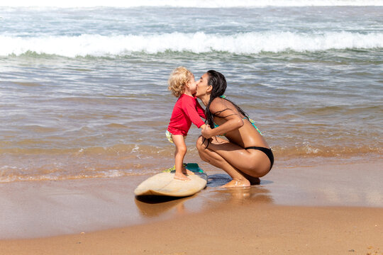 M&Atilde;E BEIJANDO FILHO NA PRAIA ENQUANTO ELE BRICA COM UMA PRANCHA DE SURF.