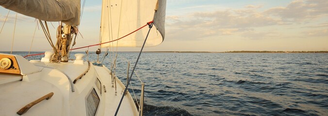 White sloop rigged yacht sailing in the sea at sunset. Clear sky. A view from the deck to the bow,...