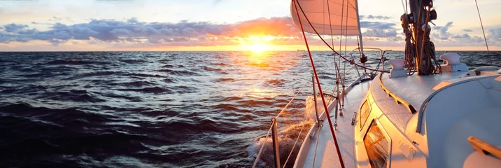Rolgordijnen Jacht zeilen in een open zee bij zonsondergang. Vergrote weergave van het dek, de mast en de zeilen. Heldere lucht na de regen, dramatische gloeiende wolken, gouden zonlicht, golven en waterspatten, cycloon. Episch zeegezicht © Aastels